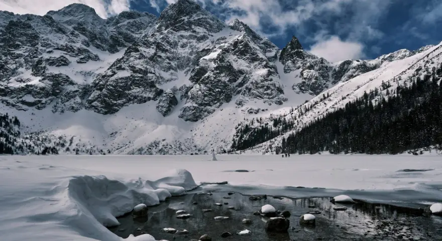 Zamarznięte Morskie Oko, otoczone ośnieżonymi szczytami Tatr. W oddali widać ludzi spacerujących po lodzie, a na pierwszym planie płynie strumyk.