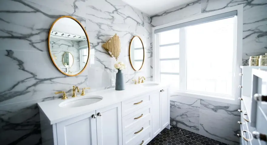 A modern bathroom vanity installation featuring white cabinets, marble walls, gold accents, and dual sinks.