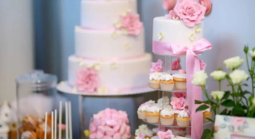 Eleganckie, białe torty weselne i babeczki ozdobione różowymi kwiatami i wstążkami. Idealne na słodki stół, inspiracja jakie ciasta na wesele.