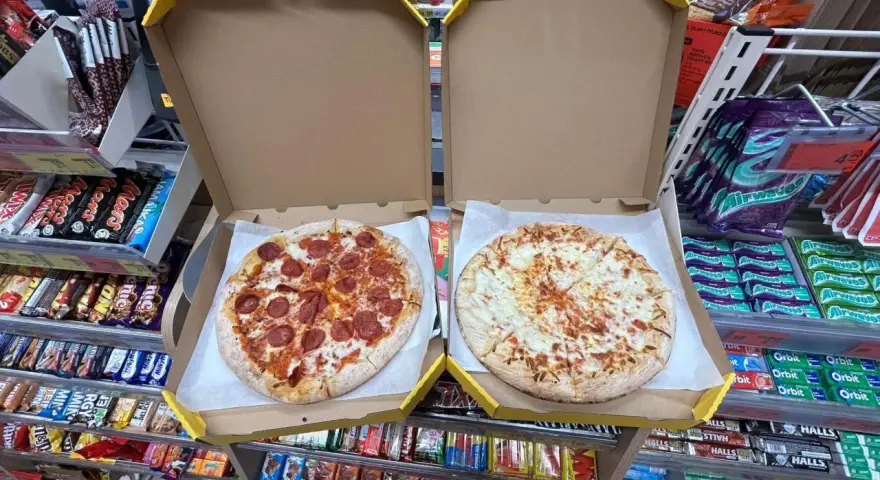 Dwie pizze w pudełkach, jedna z salami, druga serowa. Obok półki ze słodyczami i napojami w sklepie Żabka.