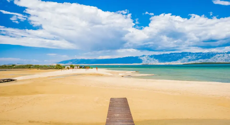 Drewniany pomost prowadzi na piaszczystą plażę Ninskiej laguny. W tle turkusowe morze i góry.