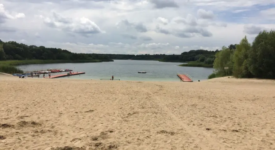 Piaskowa plaża nad jeziorem, z pomostami i łódkami. W oddali las, chmury na niebie. Idealne miejsce na wypoczynek nad wodą.