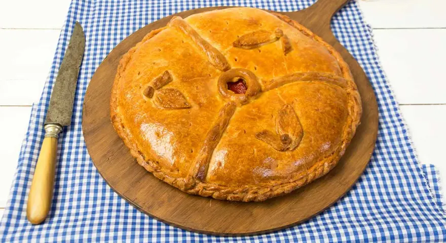 Empanada dorada y decorada sobre tabla de madera, con cuchillo y mantel a cuadros.