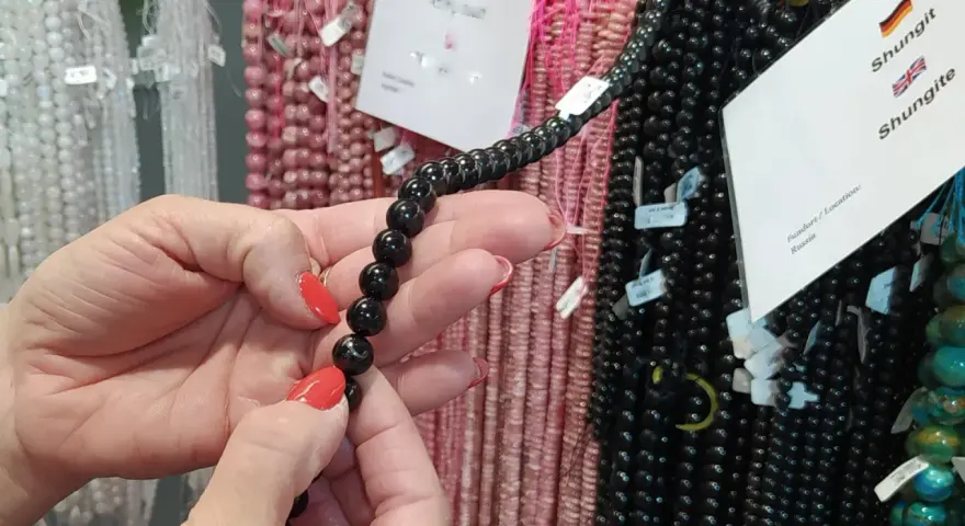 Manos con uñas rojas sostienen un collar de cuentas negras de shungita. En el fondo, se ven hilos de cuentas rosas y blancas.