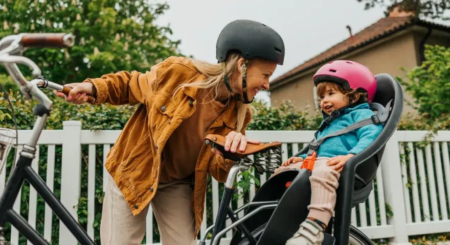 En kvinna med hjälm ler mot ett litet barn i en cykelbarnstol. De är redo att cykla med bebis i det vackra vädret.