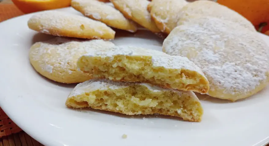 Biscotti morbidi all'arancia spolverati di zucchero a velo su un piatto bianco, con arance fresche sullo sfondo.
