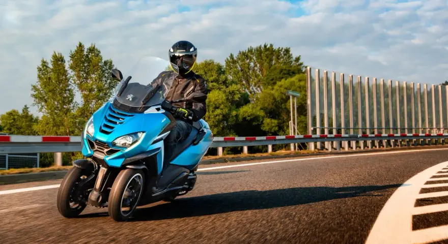 Niebieski Peugeot Metropolis 400 z kierowcą w kasku pędzi po zakręcie drogi, otoczony zielenią i błękitnym niebem.