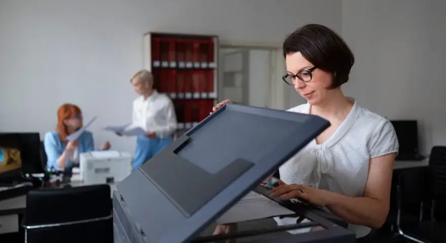 Frau mit Brille bedient einen Drucker, vielleicht um einen Epson Drucker zu installieren. Im Hintergrund arbeiten Kolleginnen.