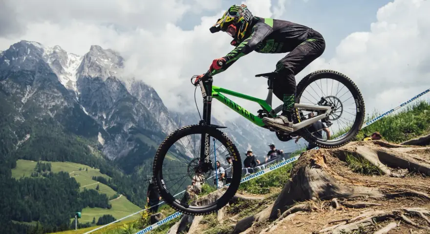 Troy Brosnan in mid-air on a green downhill bike, with a dramatic mountain backdrop.