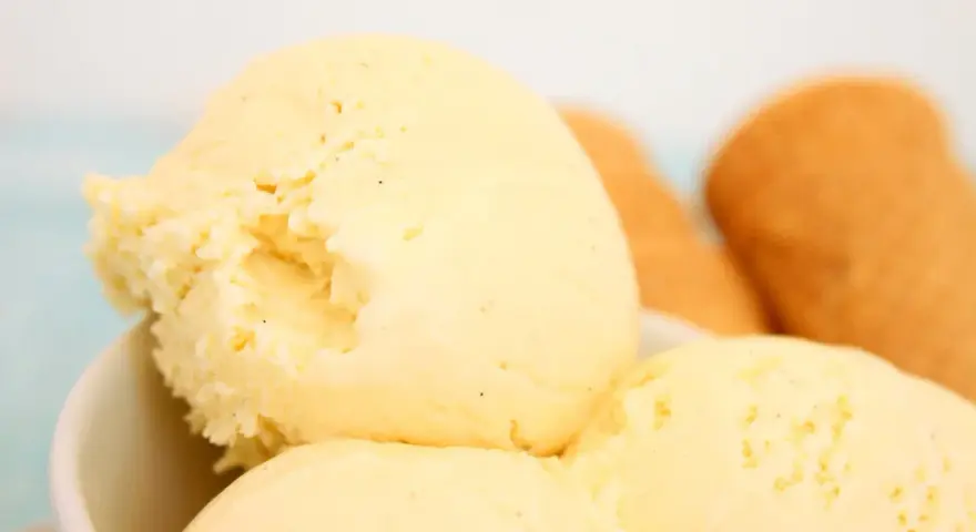 Cremoso helado de vainilla con motas oscuras, servido en un tazón blanco. Al fondo, conos de helado dorados esperan.