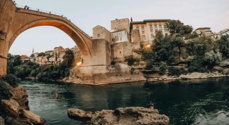 Iconic Stari Most bridge over the Neretva River, a must-see for any Bosnia travel adventure. People watch from the bridge as swimmers enjoy the emerald waters below.