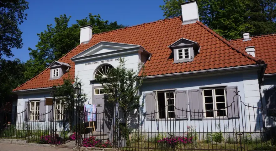 Jasnoniebieski dworek Sierakowskich z czerwoną dachówką, otoczony zielenią i ogrodzeniem.