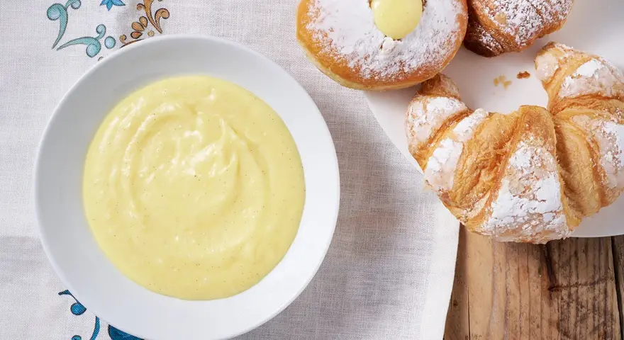 Una ciotola di soffice crema pasticcera senza uova accanto a cornetti e bomboloni spolverati di zucchero a velo.