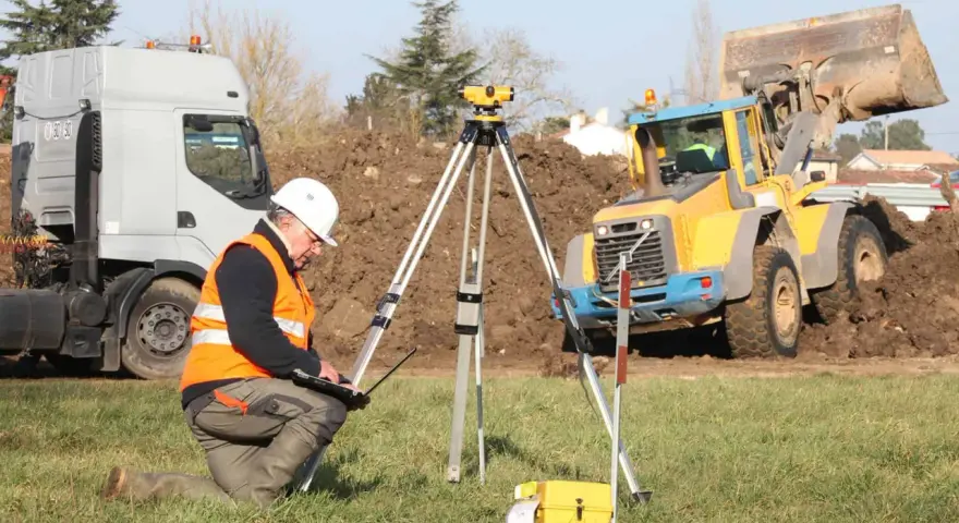 Geodeta w kamizelce odblaskowej nadzoruje odwierty geologiczne pod budowę domu. W tle koparka i ciężarówka.