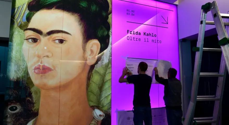 Un portrait de Frida Kahlo, avec deux hommes installant une affiche pour une exposition sur son histoire.