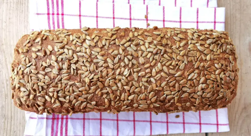 Ein rustikales Brot, bedeckt mit vielen Sonnenblumenkernen, liegt auf einem karierten Küchentuch.