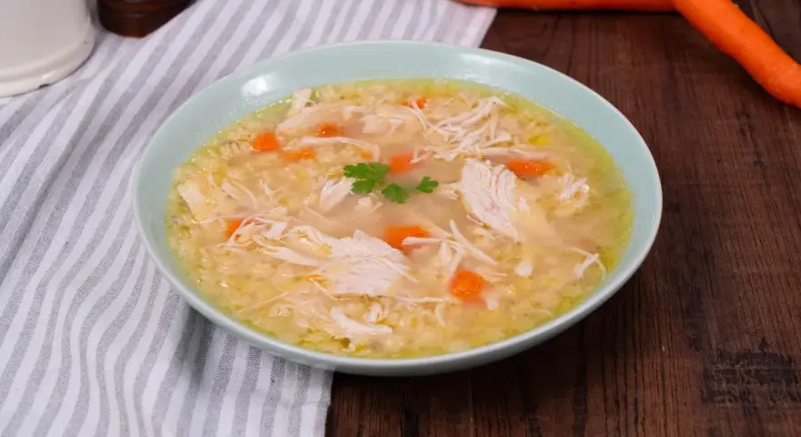 Un confortevole brodo di pollo con pezzi di pollo sfilacciato, carote e un rametto di prezzemolo, servito in una ciotola azzurra.
