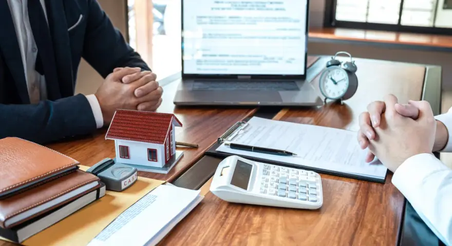 Réunion pour discuter de la faute du mandataire de gestion locative : maison modèle, calculatrice, documents et ordinateur portable sur un bureau en bois.