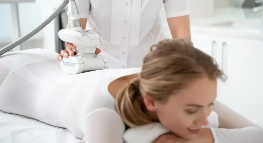 Kobieta w białym stroju poddaje się zabiegowi endermologii LPG. Widoczne są ręce terapeuty trzymające głowicę urządzenia, które masuje nogę pacjentki. Efekty zabiegu są obiecujące.
