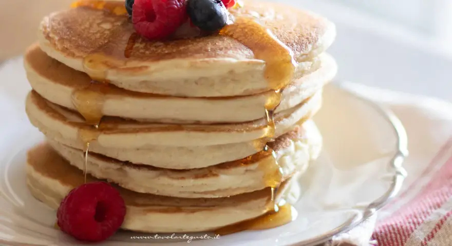 Pancakes senza uova dorati, impilati e inzuppati di sciroppo, guarniti con lamponi e mirtilli freschi.