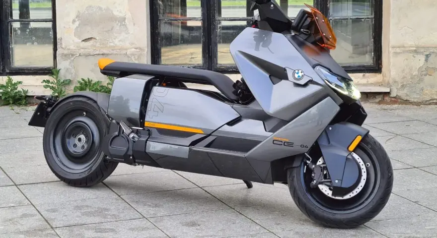 Szary skuter elektryczny BMW CE 04 z pomarańczowymi akcentami stoi na bruku przed budynkiem z dużymi oknami.