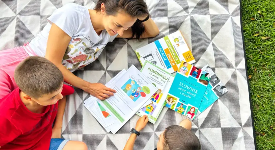 Kobieta z dwoma chłopcami leży na kocu i przegląda materiały edukacyjne o komunikacji i emocjach.
