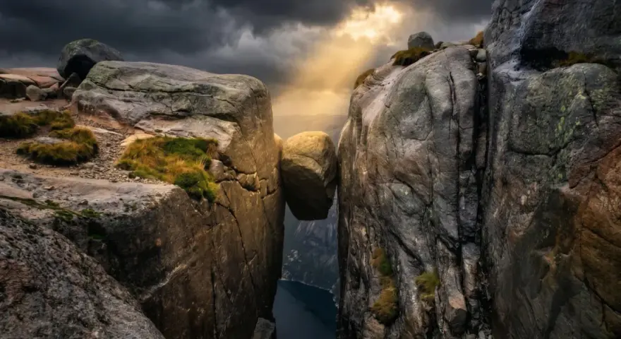 Enorm sten balanserar mellan klippor i Norge. Dramatiskt ljus bryter igenom molnen över en djup fjord.