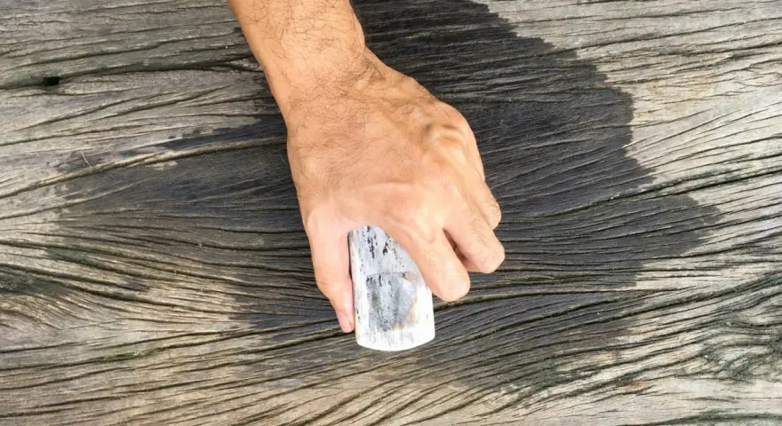 En hand håller en borste för att skura altanen. Träet är väderbitet och har en mörk, fuktig yta där borsten nyss varit.