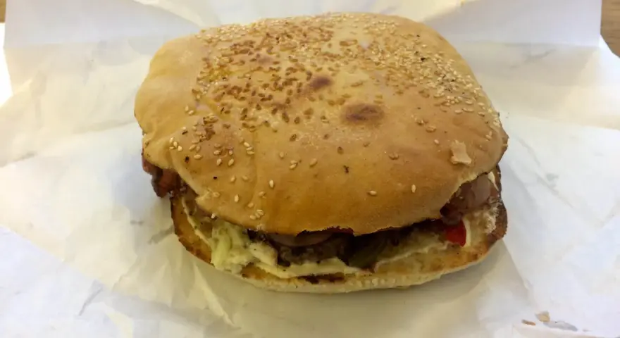 Burger z sezamem na białym papierze, widoczne składniki: mięso, sos, warzywa.