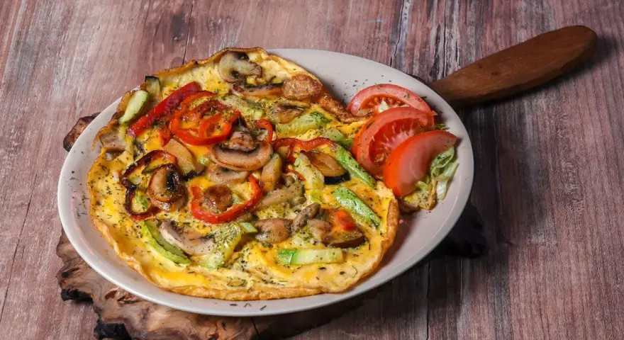 Pyszny omlet z pieczarkami, papryką i cukinią, udekorowany pomidorem i sałatą.