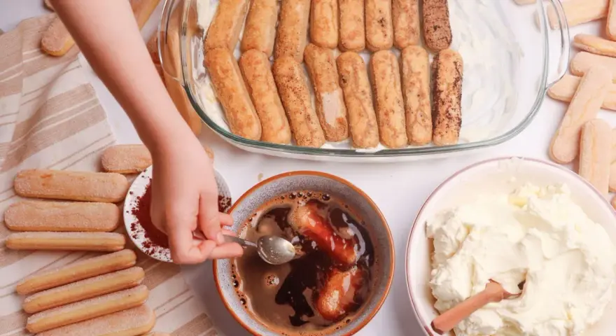 Dziecko zanurza biszkopty w kawie, przygotowując warstwy do tiramisu. Obok krem i kakao.