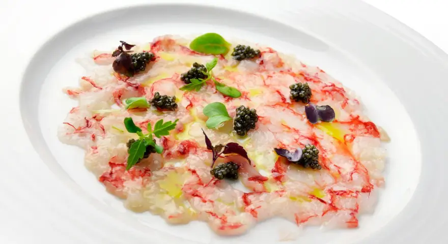 Delicado carpaccio de gambas con caviar negro, aceite de oliva y brotes verdes, presentado en un plato blanco.