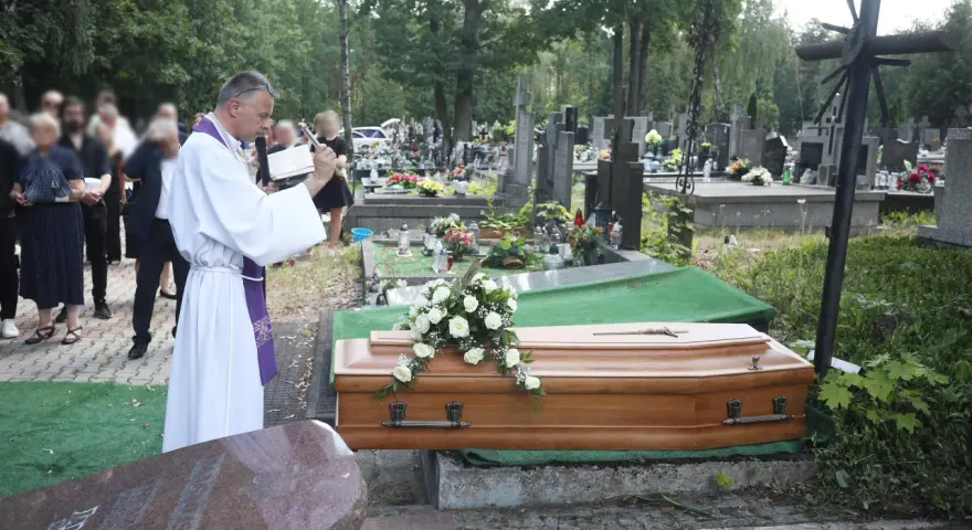 Wojciech Magnuski żegna zmarłego na cmentarzu. Ksiądz w białej szacie czyta z księgi przy trumnie ozdobionej białymi różami.