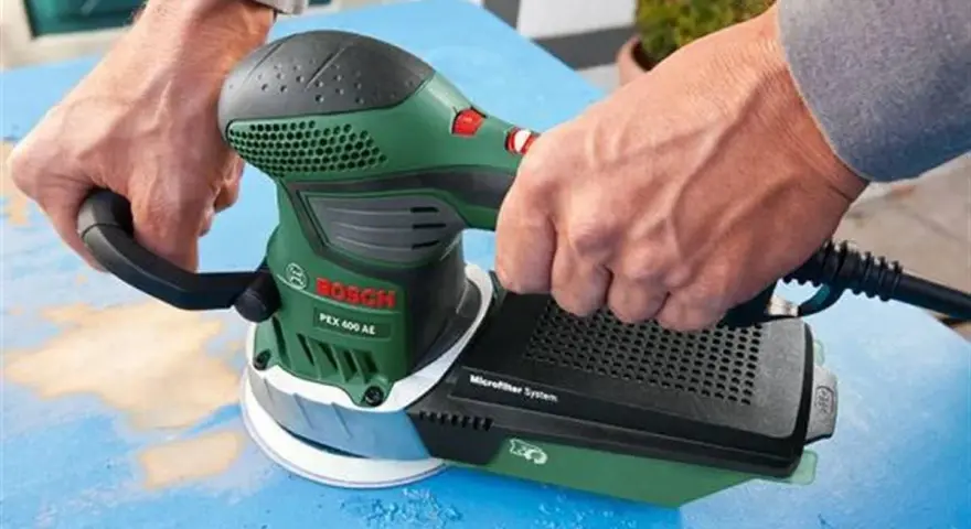 Dłonie trzymają zieloną szlifierkę Bosch PEX 400 AE. To szlifierka oscylacyjna a mimośrodowa, która usuwa starą farbę z niebieskiej powierzchni.