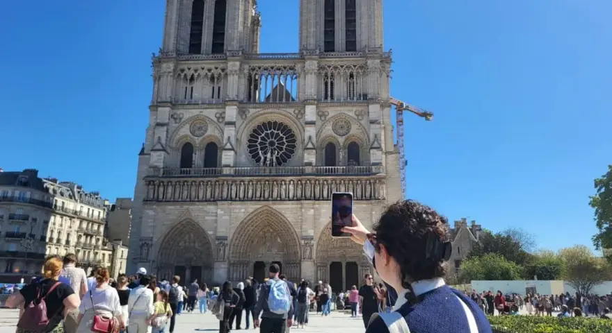 Una persona toma una foto de la fachada de Notre Dame. Mucha gente pasea por la plaza, disfrutando de su visita a Notre Dame.
