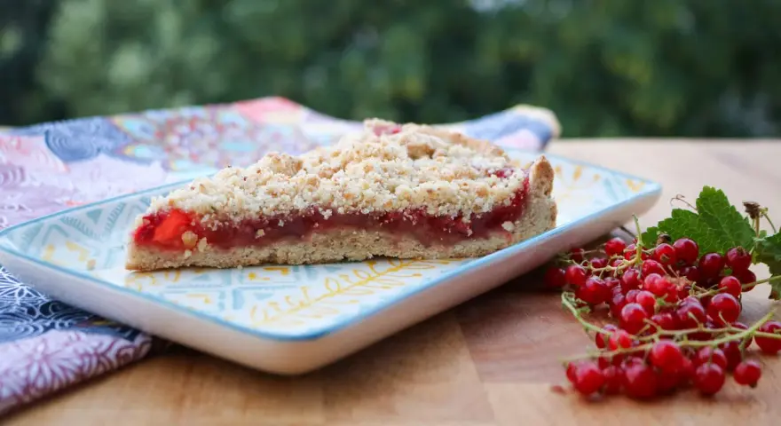 Ein Stück saftiger Johannisbeer Streuselkuchen mit frischen roten Johannisbeeren daneben.