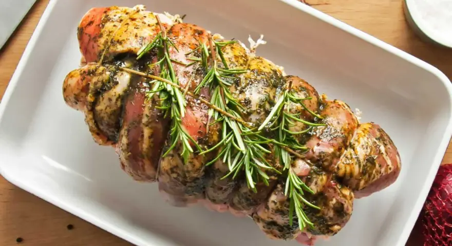 Rôti de porc Orloff, assaisonné et garni de romarin frais, prêt à être cuit dans un plat blanc.