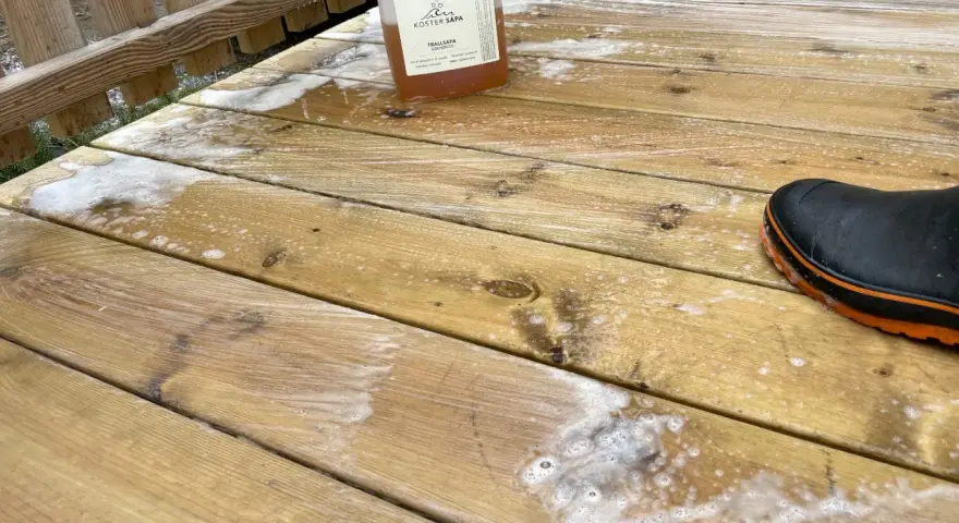 En flaska linoljesåpa står på en altan som håller på att rengöras. En svart stövel med orange kant syns i nedre högra hörnet.