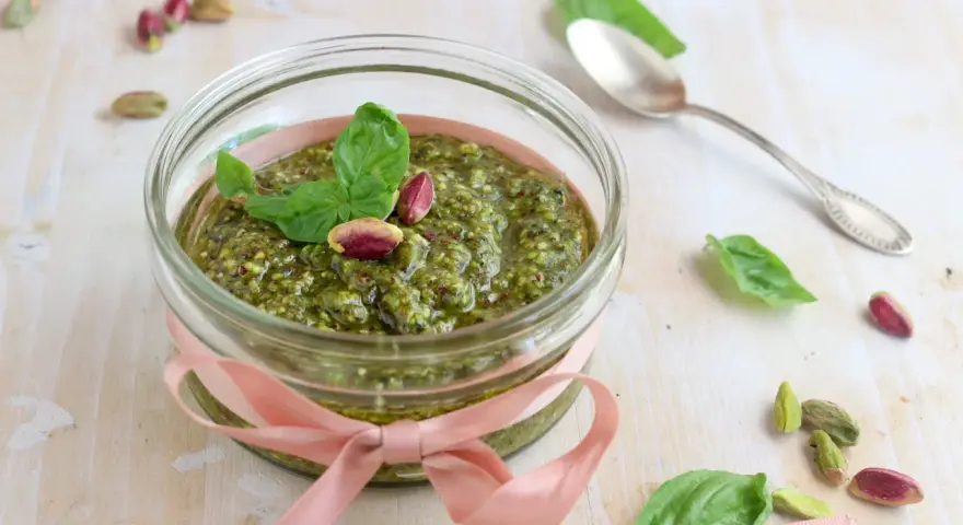 Un vasetto di pesto di pistacchi, decorato con un nastro rosa e guarnito con foglie di basilico e pistacchi interi, pronto per essere gustato.