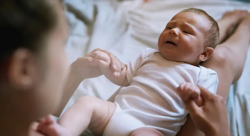 Płaczliwe niemowlę w białym body, trzymające mamę za rękę. Co robić, gdy pojawia się histeria u niemowlaka?