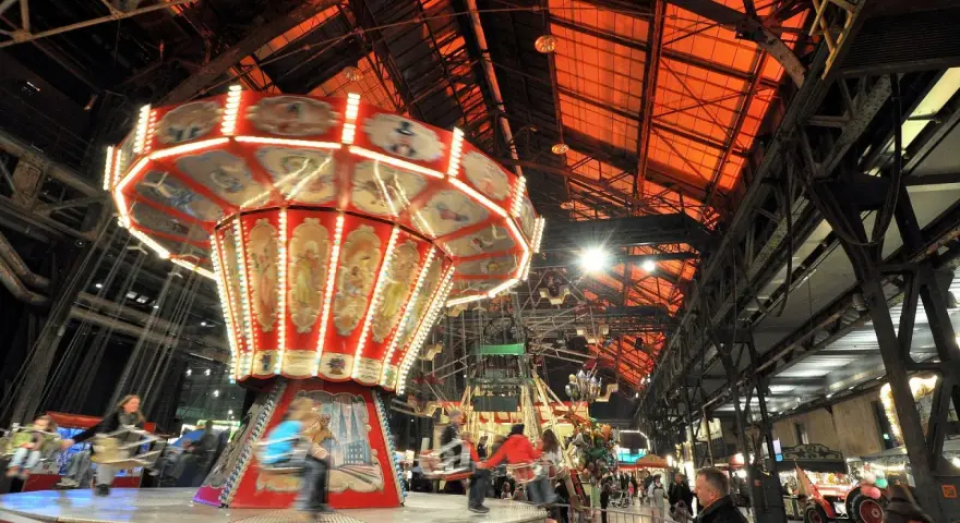 Drehende Kettenkarussells und bunte Lichter auf einem historischen Jahrmarkt in einer alten Industriehalle.