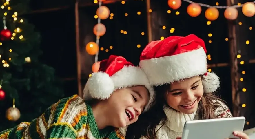 Dos niños con gorros de Santa Claus disfrutan de la tablet, felices cuando dan vacaciones de Navidad, rodeados de luces festivas y un árbol.