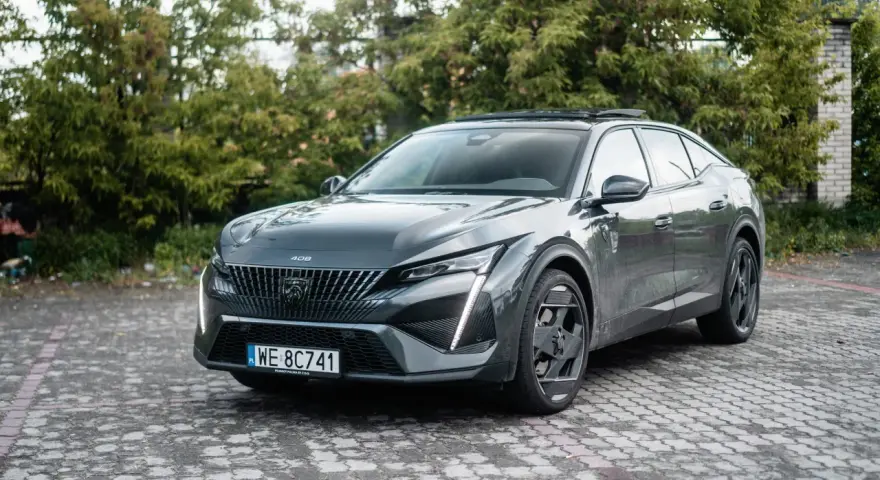 Nowoczesny Peugeot 408 Hybrid w ciemnoszarym kolorze, zaparkowany na brukowanym podjeździe, otoczony zielenią.