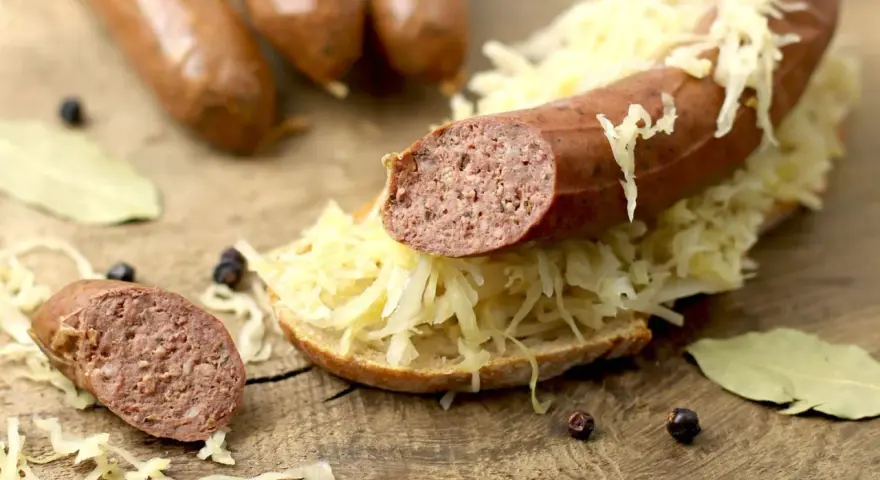 Lungenwurst zubereiten: Eine Scheibe Brot mit Sauerkraut und Lungenwurst belegt, daneben weitere Würste, Gewürze und Lorbeerblätter auf Holzuntergrund.