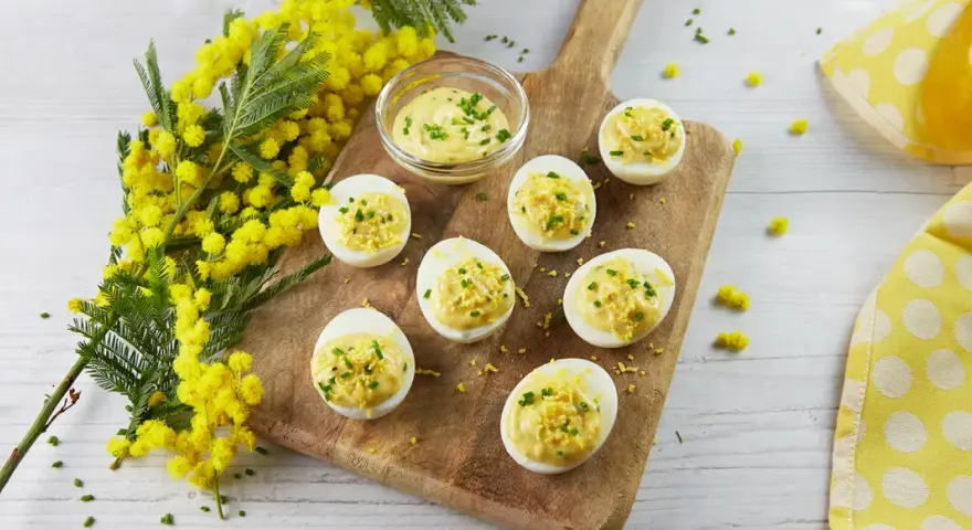 Œufs mimosa garnis d'une sauce crémeuse et de ciboulette, présentés sur une planche en bois avec des fleurs de mimosa. Une recette printanière.