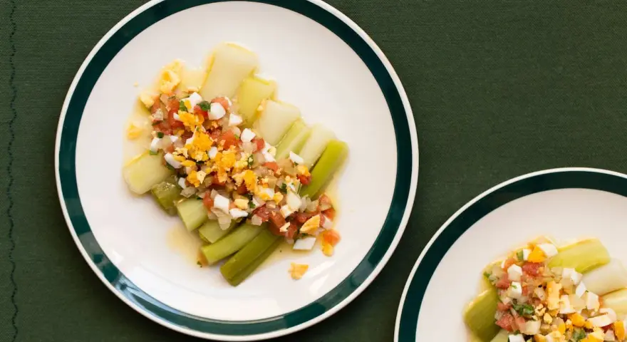 Puerros a la vinagreta servidos en platos blancos con borde verde, cubiertos con una mezcla de tomate picado, huevo duro y perejil.