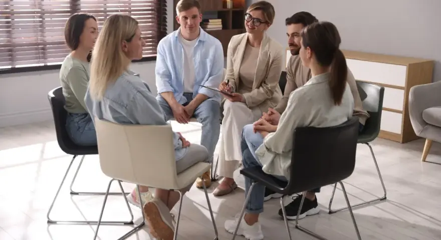 Grupa ludzi siedzi w kręgu, słuchając terapeuty. To scena z ośrodka dla uzależnionych, gdzie wsparcie i zrozumienie pomagają w powrocie do zdrowia.