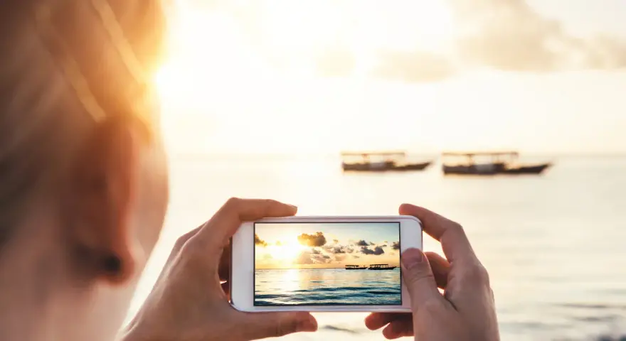 Une personne prend une photo d'un coucher de soleil sur la mer avec des bateaux au loin. Une belle photo à faire à la plage.