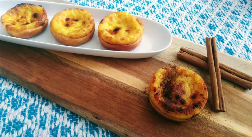 Vier goldbraune, karamellisierte Pastéis de Nata auf einem weißen Teller, daneben Zimtstangen auf einem Holzbrett.