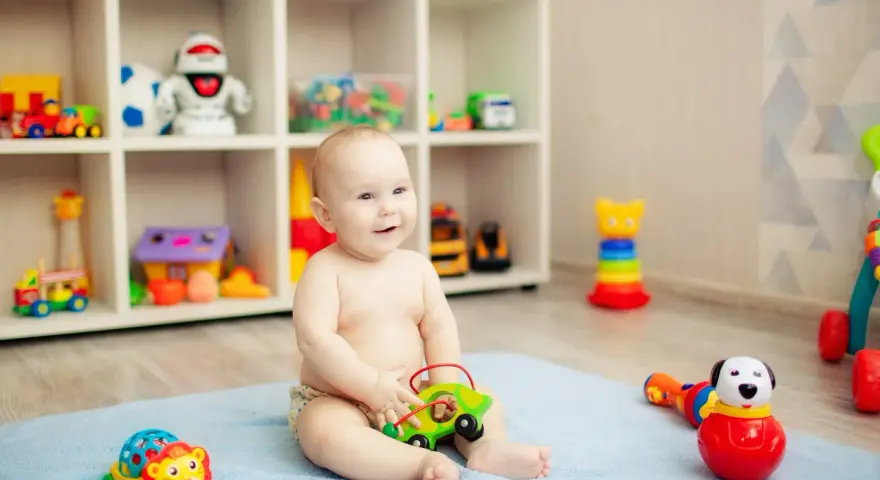 Uśmiechnięte, 8-miesięczne dziecko nie siedzi, bawi się zielonym autkiem na niebieskim kocyku. W tle regał z zabawkami.